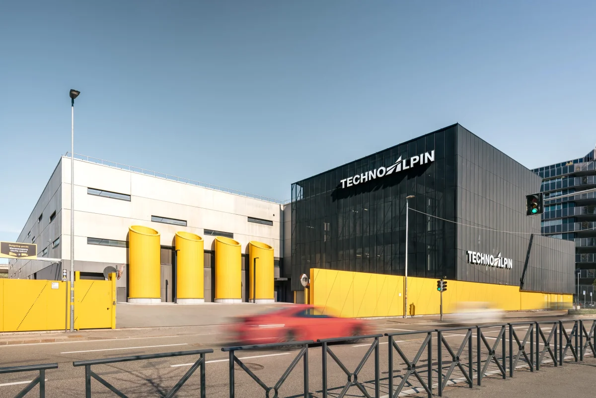 Sede produttiva della Technoalpin, azienda che si dedica alla neve artificiale. Nella foto viene mostrata la strada della zona industriale, con la scia di alcune auto in corsa. L'azienda si mostra con dei cancelli gialli e la struttura a parallelepipedo nero a destra con il logo Technoalpin e a sinistra di colore grigio con delle finestre rettangolari. Cielo è limpido.