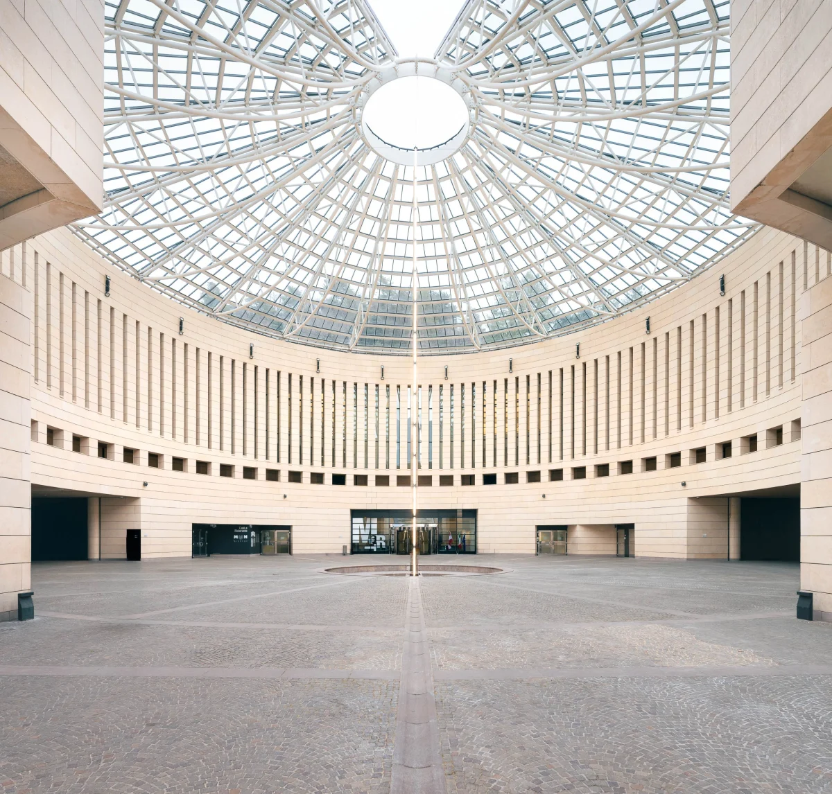 Architettura moderna: Museo MART, senza persone e in una giornata nuvolosa. Pattern di finestre verticali strette. In centro al piazzale, una colonna di luci che sale verso l'alto. La cupola è trasparente con una struttura di sostegno. Il piazzale è composto da sanpietrini