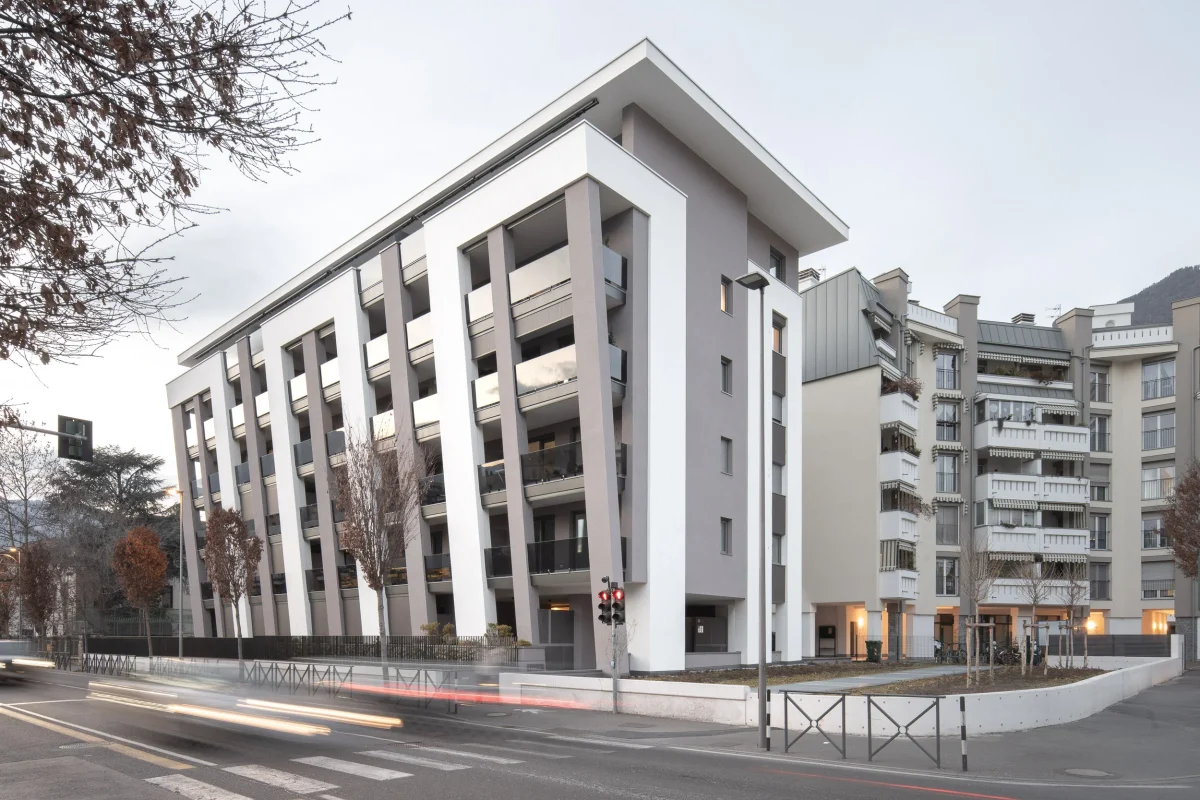 Edilizia: Palazzo Lotus (Habitat) in viale Druso a Bolzano, costituito da lastroni bianchi e grigi, + largo in altezza che alla base, come un parallelepipedo. In strada scie luminose di auto in movimento