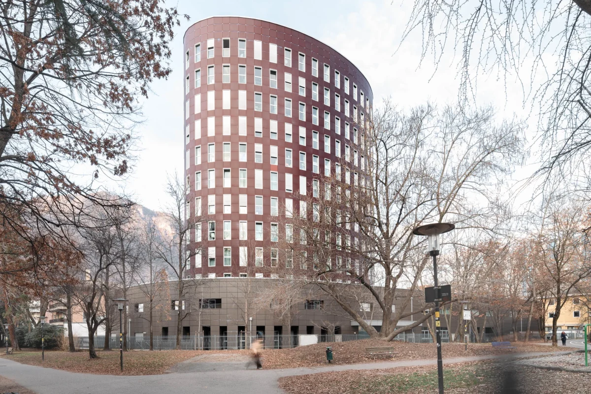 Architettura moderna: Palazzo circolare di colore bordeaux con un pattern ripetitivo di finestre, chiamato Univercity. Struttura utilizzata come residenza di studenti universitari. Il palazzo si trova in viale Europa nel quartiere Europa. Alla base della struttura ci sono persone che camminano e sono ritratte in movimento, non ben distinguibili