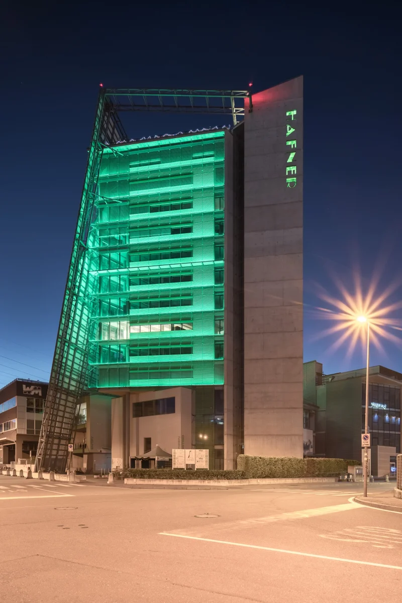 Architettura moderna: Torre Hafner a Bolzano, illuminata di luci verdi e la scritta HAFNER in verticale. Siamo al crepuscolo e la strada è illuminata
