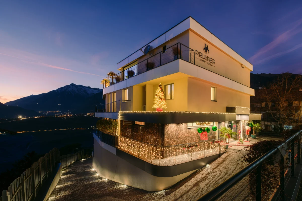 Hospitality: Prunner Luxury Suites, visione al crepuscolo di questa struttura di colore giallo con addobbi natalizi ed illuminazione. il cielo è blu con delle sfumature viola e qualche nuvola. La struttura ha 3 piani, con la parte al piano terra di color marrone scuro e addobbi natalizi.