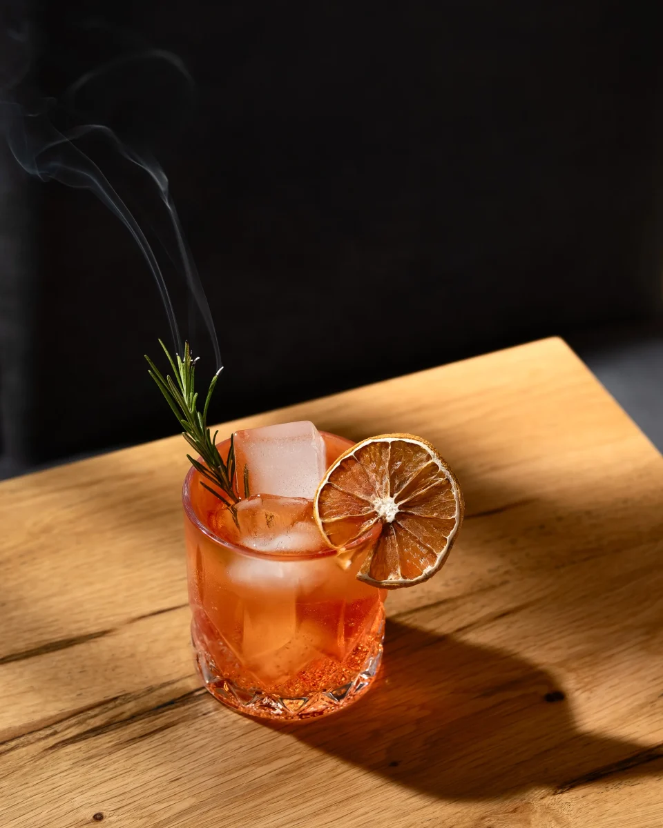 Bicchiere di aperitivo di colore rosso, con un arancio essiccato sul bicchiere, del rosmarino fumante. Su un tavolo, illuminato dal sole, tra le foglie