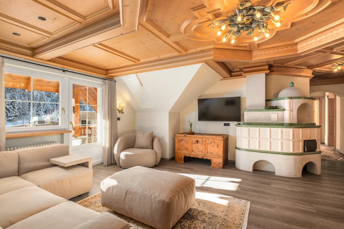 interni: Villa Leck a Selva di Val Gardena, prime. ore del mattino in questa zona giorno di un appartamento. Soffito in legno e divani di color marrone, un tappeto in centro alla stanza. Sulla destra un mobile e una tv, oltre ad una stufa ed un corridoio. Dalla finestra si vedono degli alberi innevati