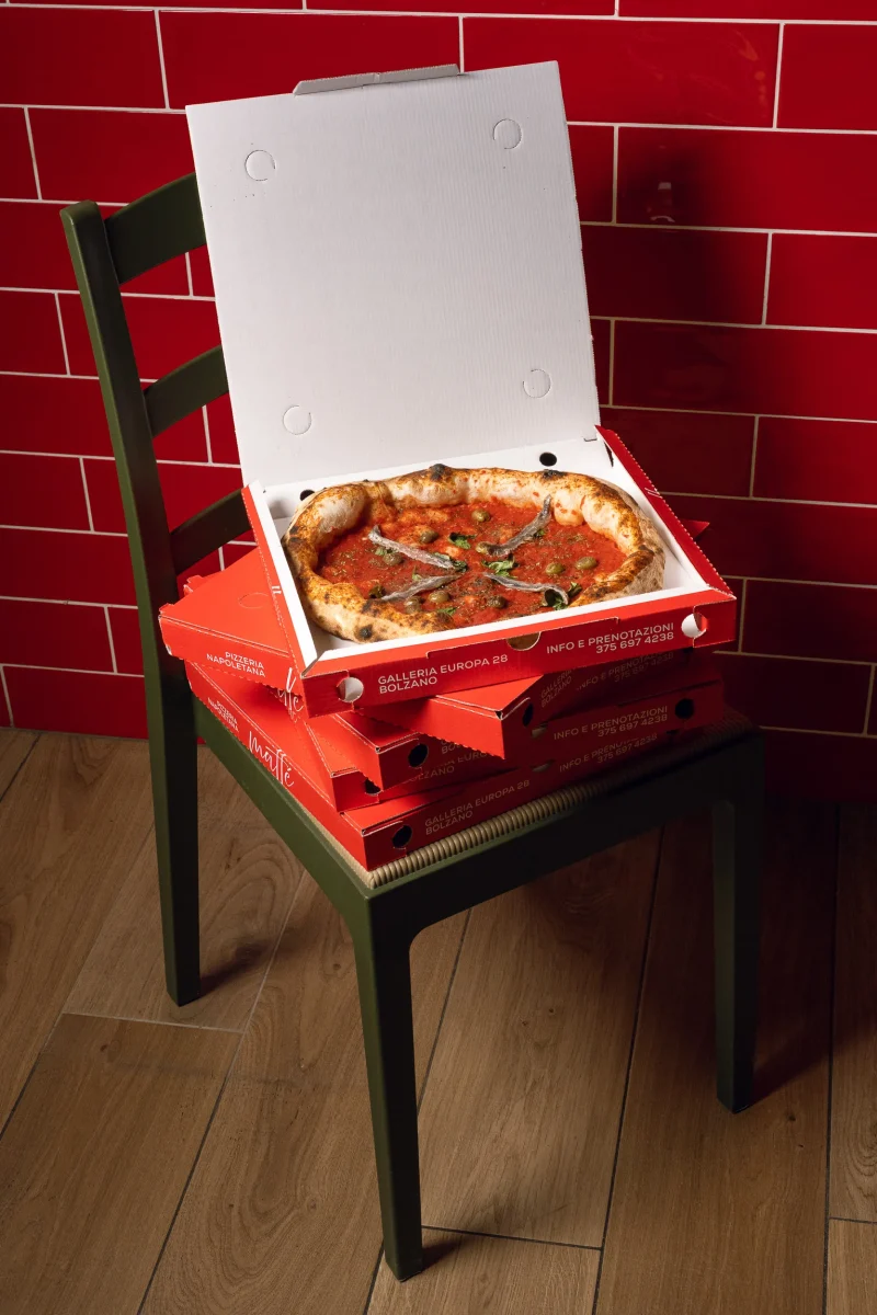 cartone di pizza d'asporto appoggiato su pila di cartoni, su una sedia e parete di mattonelle rosse
